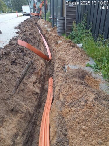 nedgravning af fibernet, hvor en smal, dyb rende er gravet langs en vej og en bygning. I renden er der lagt orange fiberbeskyttelsesrør, som endnu ikke er tildækket. Den opgravede jord ligger langs begge sider af renden, og der ses entreprenørmaskine og arbejdskøretøj i baggrunden, hvilket tydeligt indikerer aktivt anlægsarbejde. Motivet dokumenterer installationsfasen, før fibernettet dækkes til og området genetableres.