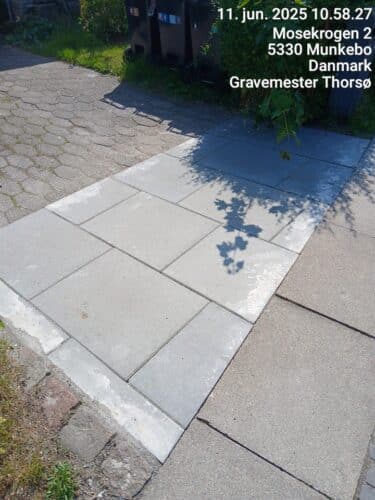 fortovs- og indgangsparti, hvor belægningen er blevet genetableret efter et gravearbejde. Der er lagt nye, lyse betonfliser i et rektangulært felt, som tydeligt adskiller sig fra den ældre omkringliggende belægning. Overgangen mellem de nye fliser og den eksisterende belægning er synlig, hvilket indikerer nyligt udført jord- eller ledningsarbejde. Området fremstår ryddet og færdiggjort, uden åbne udgravninger.