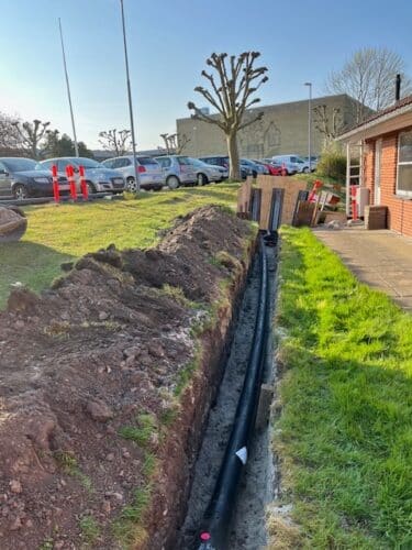 aktivt gravearbejde i forbindelse med nedgravning af fjernvarme. En lang, åben rende løber gennem et grønt areal langs en bygning. I renden er der lagt sorte fjernvarmerør, som endnu ikke er tildækket. Den opgravede jord ligger langs kanten af renden, og området er tydeligt under udførelse. Der ses afspærring og arbejdsudstyr, hvilket understreger, at arbejdet er i gang. Motivet dokumenterer selve installationsfasen før genetablering af terræn.